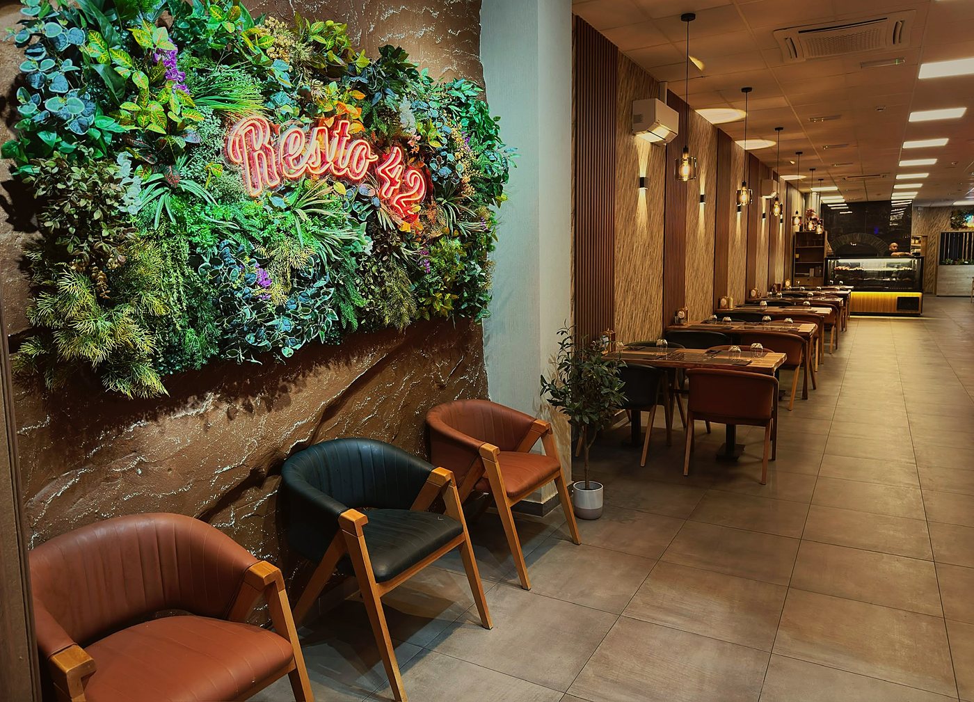 Lange zaal van Resto 42 met een plantenwand en het neonbord Resto 42, leren stoelen, notelaren tafels en achteraan de toonbank