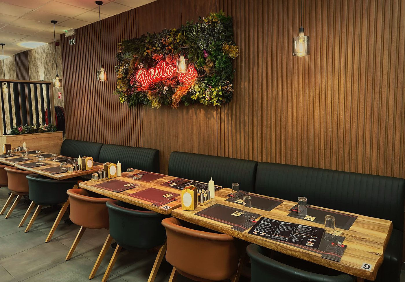 Groene zitbanken en bruine stoelen aan notelaren tafels, met houten wandlatten en een plantenwand met het neonbord Resto 42